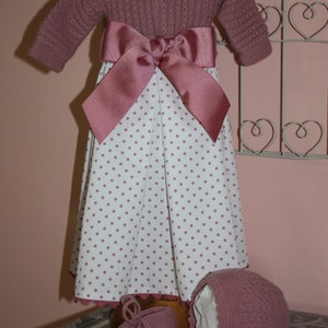 Puede incluir: Un vestido de bebé de punto rosa con una falda de lunares blancos y un gorro y zapatos a juego de punto rosa.
