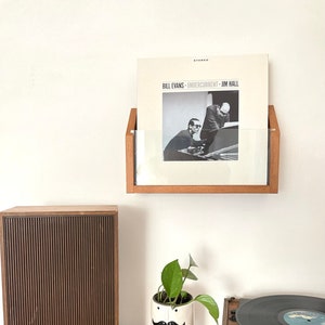 May include: A wooden record shelf with a vinyl record by Bill Evans, Undercurrent, Jim Hall. A brown speaker, a white ceramic planter with a mustache and a green plant, and a vintage record player are on a wooden surface.