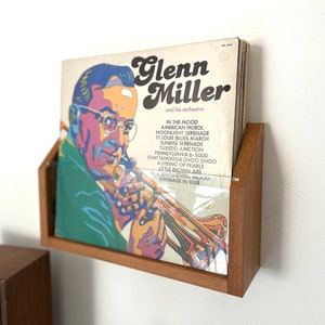 May include: A wooden wall mount record holder with a vinyl record featuring a portrait of Glenn Miller and his orchestra. The record cover features the text "Glenn Miller and his orchestra" and a list of songs including "In the Mood", "American Patrol", "Moonlight Serenade", "St. Louis Blues March", "Sunrise Serenade", "Tuxedo Junction", "Pennsylvania 6-5000", "Chattanooga Choo Choo", "A String of Pearls", "Little Brown Jug", "A Sentimental Mood", and "Serenade in Blue".