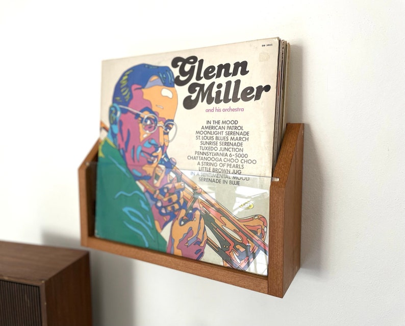 May include: A wooden wall mount record holder with a vinyl record featuring a portrait of Glenn Miller and his orchestra. The record cover features the text "Glenn Miller and his orchestra" and a list of songs including "In the Mood", "American Patrol", "Moonlight Serenade", "St. Louis Blues March", "Sunrise Serenade", "Tuxedo Junction", "Pennsylvania 6-5000", "Chattanooga Choo Choo", "A String of Pearls", "Little Brown Jug", "A Sentimental Mood", and "Serenade in Blue".