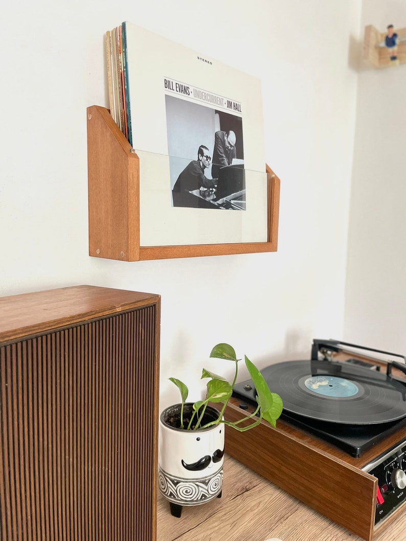 May include: A wooden record shelf with a vinyl record by Bill Evans titled "Undercurrent" with a black and white photo of two men playing music. The shelf is mounted on a white wall. A turntable, a potted plant, and a speaker are also visible in the image.