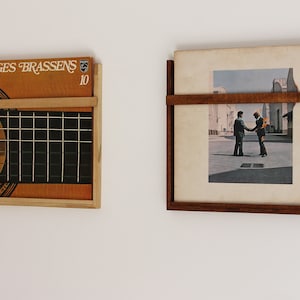 May include: Two vinyl record storage shelves mounted on a white wall. The shelf on the left is made of wood and has a metal grid. The shelf on the right is made of wood and has a single horizontal shelf. The record on the left shelf has the text "GEORGES BRASSENS 10" on the cover. The record on the right shelf has a black and white image of two men shaking hands.