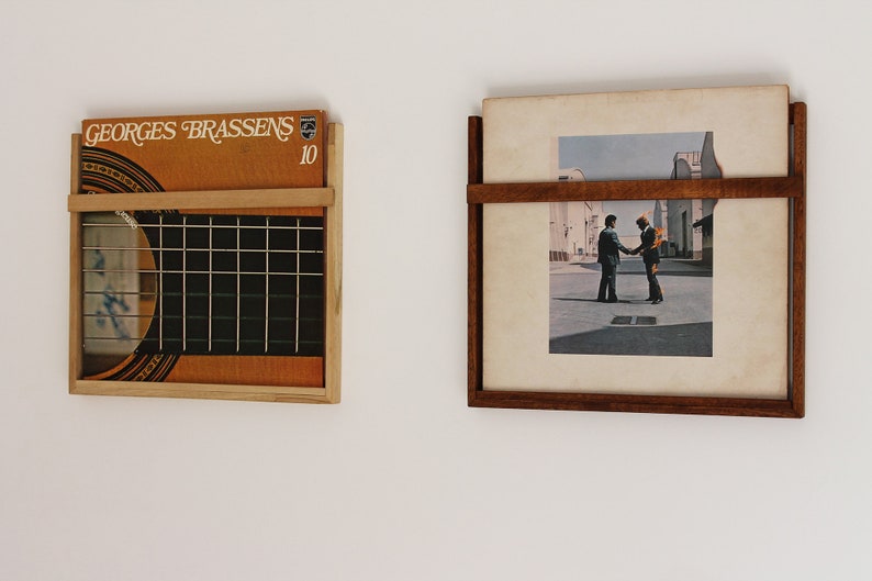 May include: Two vinyl record storage shelves mounted on a white wall. The shelf on the left is made of wood and has a metal grid. The shelf on the right is made of wood and has a single horizontal shelf. The record on the left shelf has the text "GEORGES BRASSENS 10" on the cover. The record on the right shelf has a black and white image of two men shaking hands.