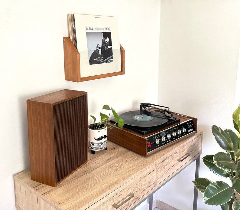 May include: A vintage record player with a wooden cabinet and a turntable. The record player is on a wooden shelf with a speaker and a record album cover on a shelf above it.