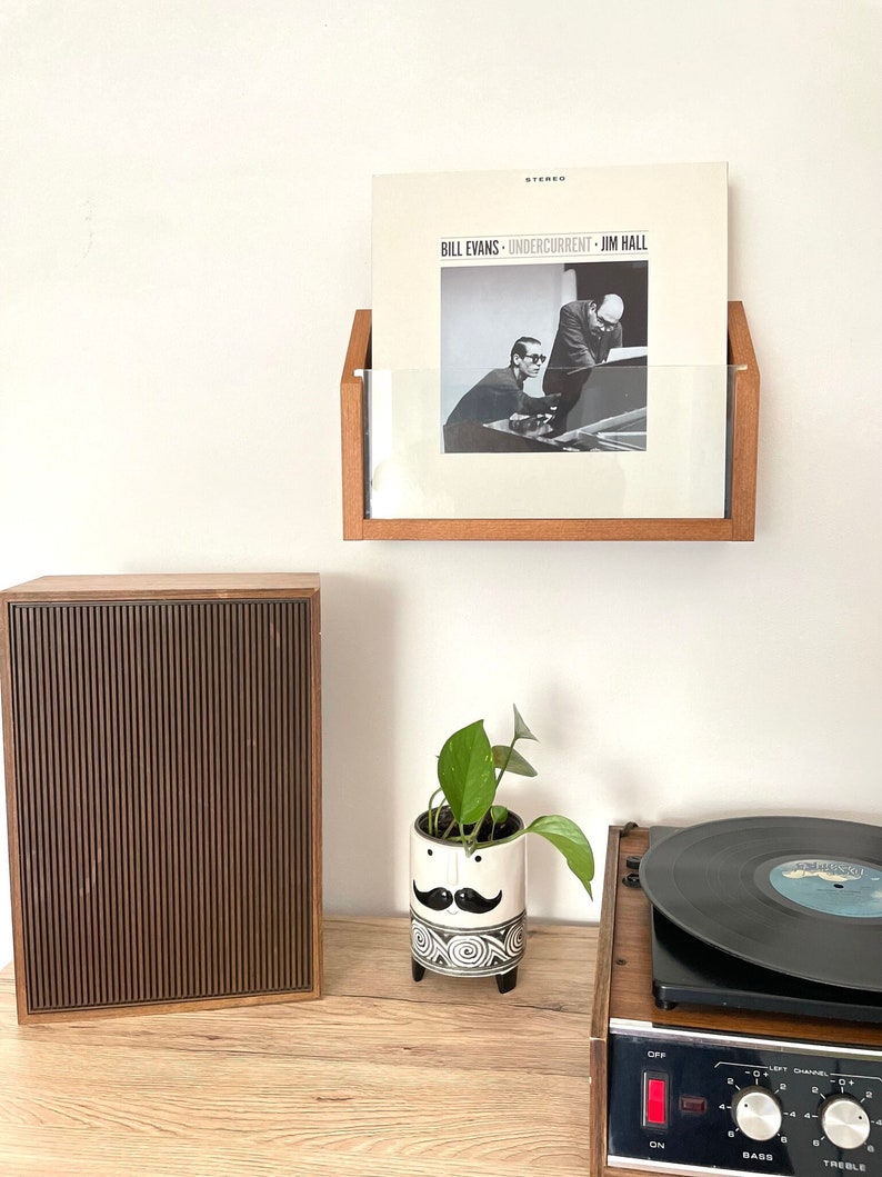 May include: A wooden record shelf with a vinyl record by Bill Evans, Undercurrent, Jim Hall. A brown speaker, a white ceramic planter with a mustache and a green plant, and a vintage record player are on a wooden surface.