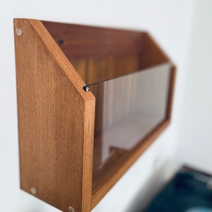 May include: A wooden wall-mounted record storage shelf with a clear acrylic front panel. The shelf is stained a light brown color and has a triangular shape.