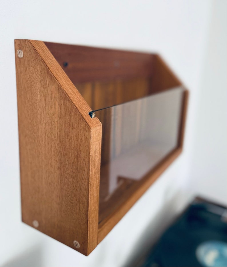 May include: A wooden wall-mounted record storage shelf with a clear acrylic front panel. The shelf is stained a light brown color and has a triangular shape.