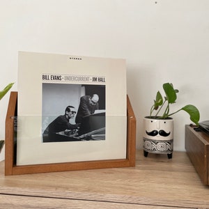 May include: A wooden record stand with a clear plastic insert holds a vinyl record by Bill Evans, "Undercurrent", featuring Jim Hall. The record stand is on a wooden surface next to a potted plant and a record player.