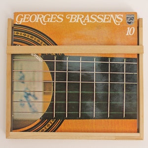 May include: A wooden record holder with a vinyl record inside. The record is by Georges Brassens and is a Philips recording. The record cover is orange and brown with a guitar on the front. The record number is 10.