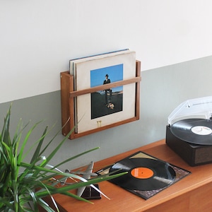 May include: A wooden wall-mounted record rack with vinyl records, a turntable with a clear dust cover, and a record on a wooden table.
