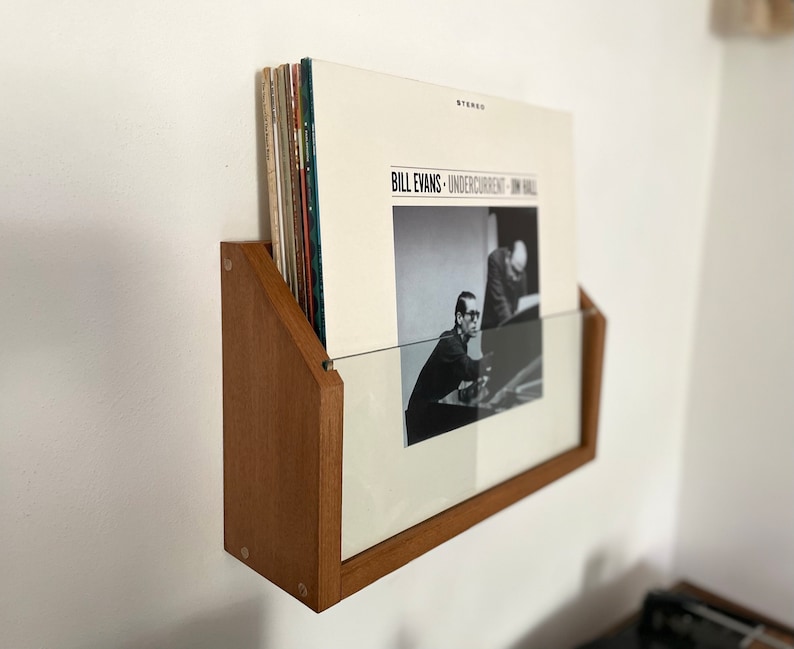 May include: A wooden wall-mounted record shelf with a clear acrylic front. The shelf is holding a stack of vinyl records, including one with the title "Bill Evans - Undercurrent - Jim Hall".