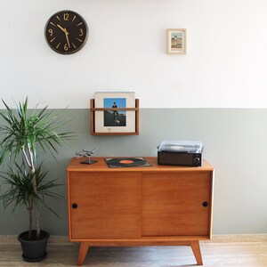 May include: A mid-century modern wooden cabinet with a sliding door, a record player, and a record rack on the wall above. A houseplant is in a black pot on the floor. A clock with black hands and gold numbers is on the wall.