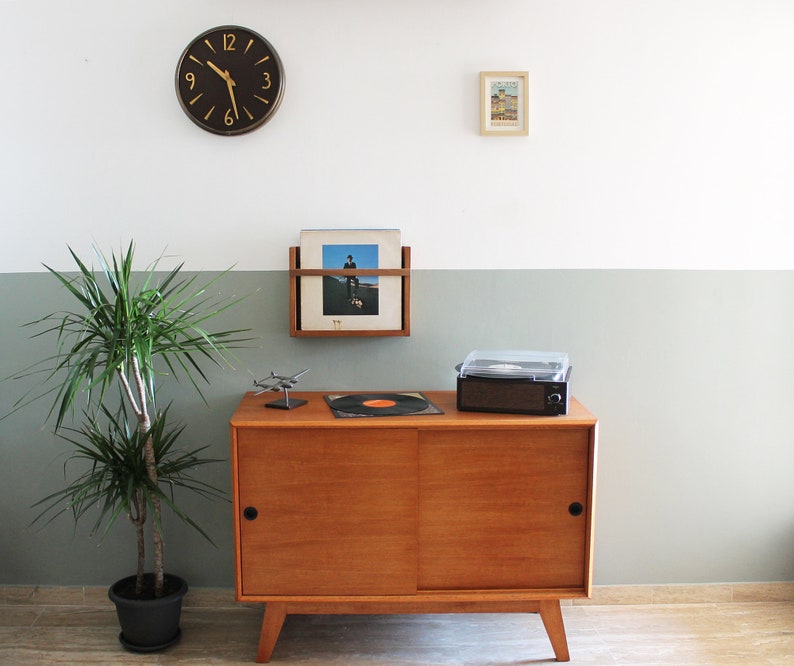 May include: A mid-century modern wooden cabinet with a sliding door, a record player, and a record rack on the wall above. A houseplant is in a black pot on the floor. A clock with black hands and gold numbers is on the wall.