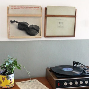 May include: Two framed vinyl records on a white wall, one with a violin on the cover and the text "HANDEL CONCERTO FOR VIOLIN - B MINOR", the other with the text "AMELIA DREXLER", a record player on a wooden table with a plant in a yellow pot with a face on it, and a sheet of music on the table.