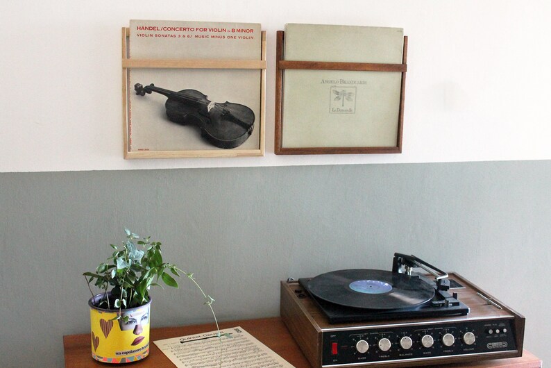 May include: Two framed vinyl records on a white wall, one with a violin on the cover and the text "HANDEL CONCERTO FOR VIOLIN - B MINOR", the other with the text "AMELIA DREXLER", a record player on a wooden table with a plant in a yellow pot with a face on it, and a sheet of music on the table.