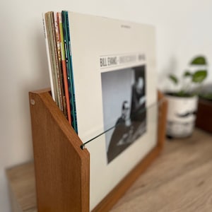 May include: A wooden record stand with a glass front, holding a stack of vinyl records. The top record features a black and white photo of Bill Evans and the text "Bill Evans - Sunday at the Village Vanguard".