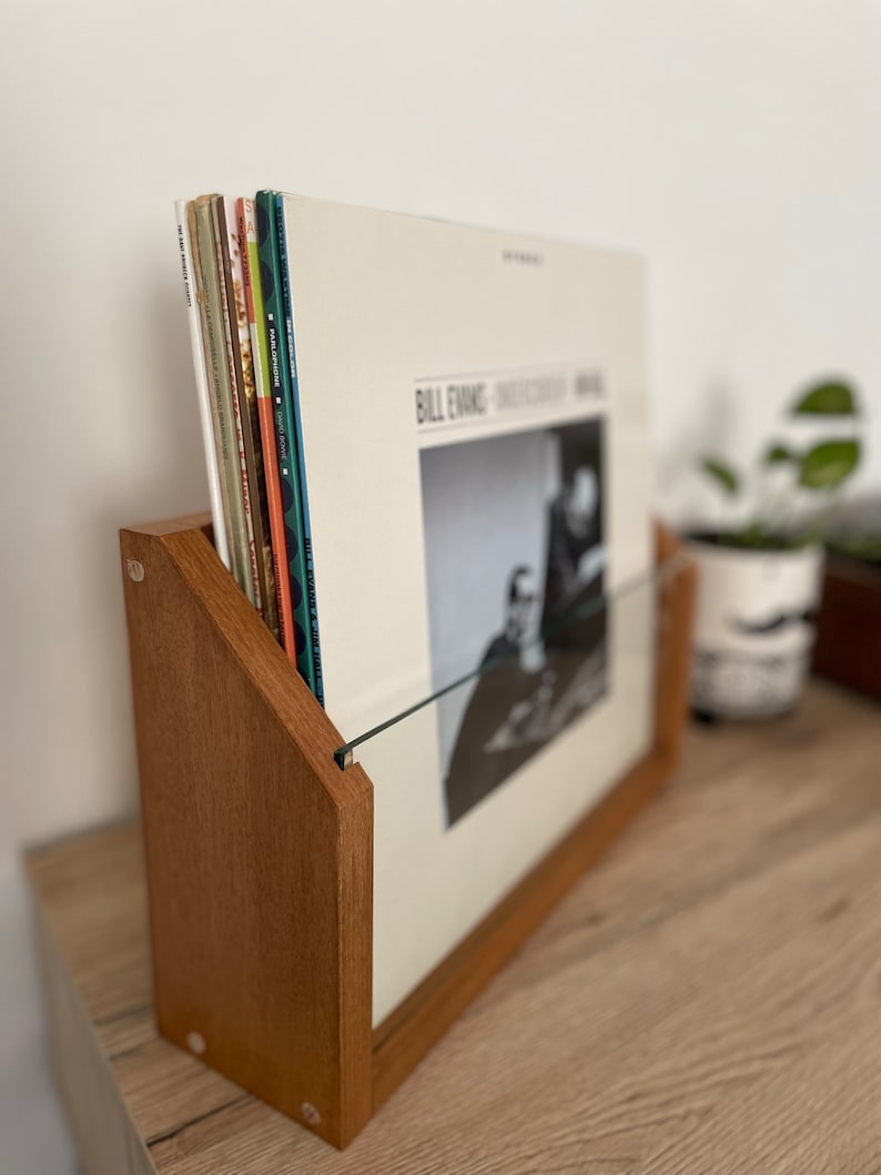 May include: A wooden record stand with a glass front, holding a stack of vinyl records. The top record features a black and white photo of Bill Evans and the text "Bill Evans - Sunday at the Village Vanguard".