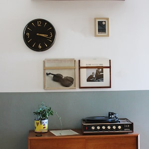 May include: A vintage record player with a wooden cabinet and a black turntable sits on a wooden shelf. A clock with black hands and gold numbers is on the wall above the record player. There are two framed album covers on the wall, one with a violin and the other with a band. The wall is painted in two shades of gray.
