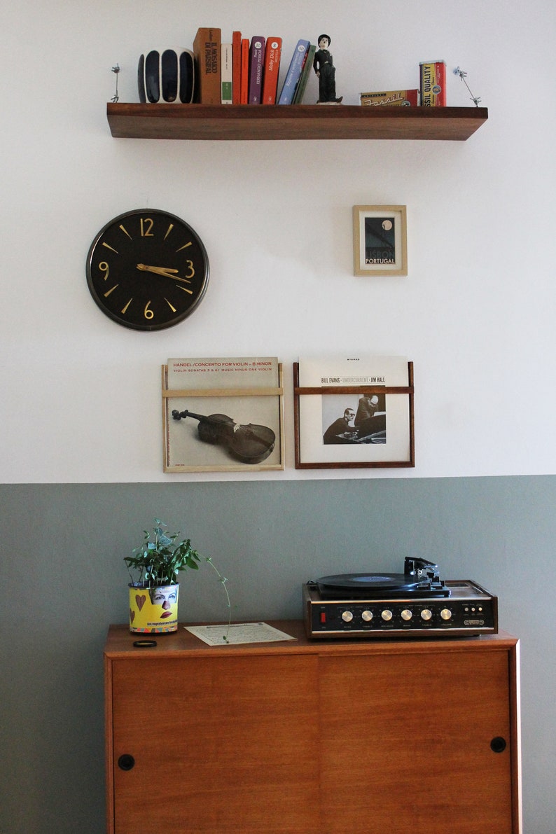 May include: A vintage record player with a wooden cabinet and a black turntable sits on a wooden shelf. A clock with black hands and gold numbers is on the wall above the record player. There are two framed album covers on the wall, one with a violin and the other with a band. The wall is painted in two shades of gray.