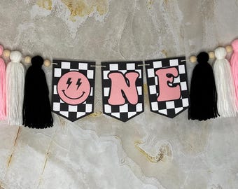 May include: Pink and black checkered banner with the word "ONE" in pink letters and a pink smiley face with lightning bolts. The banner is strung with pink, white, and black tassels.