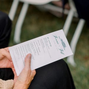 Puede incluir: Un programa de ceremonia de boda sostenido por una persona. El programa es blanco con texto verde, incluyendo los nombres "Jennie & Jeff" y los detalles de la ceremonia. La persona lleva ropa negra.