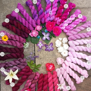 May include: A collection of 18 skeins of yarn in shades of pink, purple, and red arranged in a circle. The yarn is hand-dyed and has a gradient effect. The skeins are numbered 1 through 18.