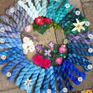 May include: A collection of 18 skeins of yarn in shades of blue, teal, and green arranged in a circle on a stone surface. The yarn is arranged in a gradient from light blue to dark green. Each skein is labeled with a number from 1 to 18.