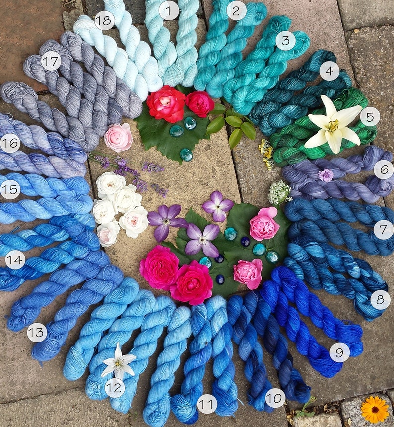 May include: A collection of 18 skeins of yarn in shades of blue, teal, and green arranged in a circle on a stone surface. The yarn is arranged in a gradient from light blue to dark green. Each skein is labeled with a number from 1 to 18.