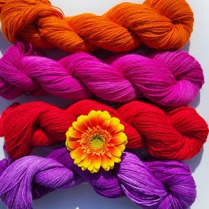 May include: Four skeins of yarn in different colors: orange, pink, red, and purple. A single orange gerbera daisy is in the center of the yarn.