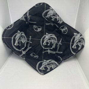 May include: Black fabric with a repeating pattern of a wolf head and a sword. The fabric is diamond shaped.