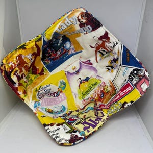May include: A square fabric bowl featuring a collage of vintage Disney movie posters. The bowl's design includes images from "Lady and the Tramp," "Cinderella," and "Snow White." The edges are lined with yellow fabric, creating a vibrant and nostalgic aesthetic.
