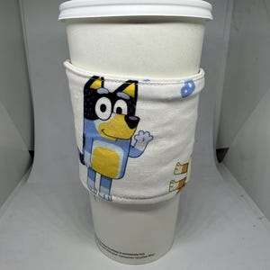 May include: A white coffee cup with a fabric sleeve featuring a cartoon dog with blue, yellow, and black markings. The sleeve has a white background with blue and white clouds. The text on the cup reads "This cup is only extremely hot.  Please use caution.  Made with post-consumer recycled fiber."