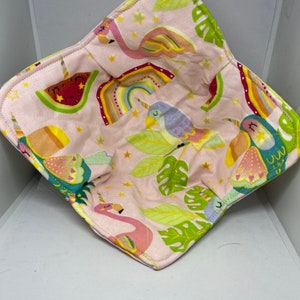May include: Pink fabric bowl with a tropical theme. The bowl features flamingos, rainbows, watermelon, and birds. The fabric is pink with yellow, green, and blue accents.
