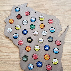 May include: A wooden cutout of the state of Wisconsin with bottle caps from various beer brands filling the outline. The bottle caps are in various colors, including red, blue, yellow, and green. The bottle caps include brands such as Bud Light, Coors, and Miller.