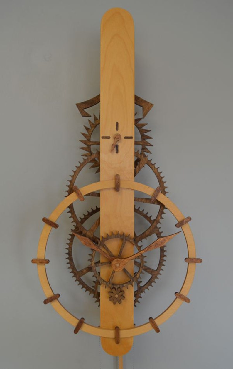 May include: Wooden wall clock with a light brown vertical base and a series of dark brown gears and a circular frame. The clock hands are made of wood. The clock has a pendulum at the bottom.