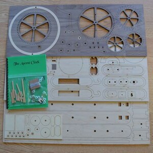 May include: Wooden clock kit with laser-cut gears and components. Includes a green booklet titled "The Ascent Clock" and a bag of hardware. The clock parts are arranged on two wooden boards, ready for assembly.