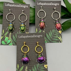 Peut inclure: Trois paires de boucles d'oreilles pendantes sur des cartes de présentation marquées LollapaloozaDesigns. Chaque paire présente un scarabée métallique suspendu à un anneau. Les scarabées affichent des teintes irisées vertes, cuivrées et violettes.