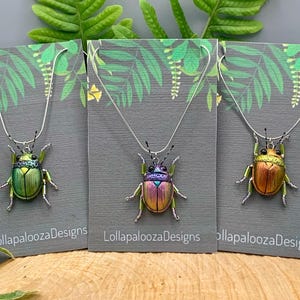 May include: Three beetle-shaped pendants on silver-tone chains, each displayed on a gray card labeled LollapaloozaDesigns. The left pendant is metallic green, the center is iridescent purple and gold, and the right is metallic copper and gold.