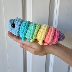 Könnte beinhalten: Ein handgefertigtes, plüschiges Raupenspielzeug in Regenbogenfarben. Die Raupe besteht aus weichem, strukturiertem Garn und hat einen blauen Kopf mit einem schwarzen Auge. Der Körper wechselt durch Türkis, Grün, Gelb, Rosa und Lila.