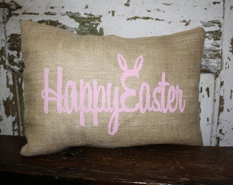 May include: A rectangular burlap cushion with the words "Happy Easter" in pink script, and a pink bunny ears graphic. The cushion is on a dark wooden surface.