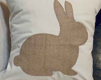 May include: A square, cream-coloured cushion featuring a burlap rabbit silhouette. The rabbit is a light brown colour and is centrally placed. The cushion is likely for decorative use.