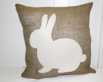 May include: A square burlap cushion featuring a large white rabbit silhouette. The cushion has a natural brown colour and a textured surface. The rabbit is centered and has long ears and a round body. Ideal for Easter.