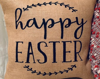 May include: Square burlap cushion with the words "happy EASTER" in dark blue script. The text is framed by a dark blue leafy design. The cushion is on a white surface.