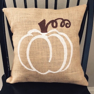 May include: A square burlap pillow with a white pumpkin design. The pumpkin outline is painted on the pillow's front, with a brown stem and curly vine. The pillow is sitting on a black chair.