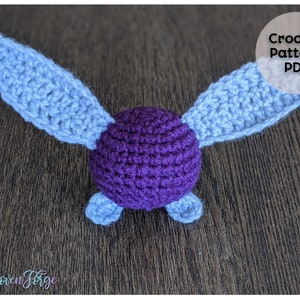 Puede incluir: Patrón de crochet PDF para un juguete amigurumi morado y azul claro con alas. El juguete tiene forma de pájaro o criatura voladora.