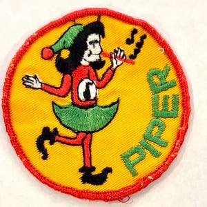 May include: A circular embroidered patch with a yellow background and a red border. It features a cartoon character wearing a green hat, red shirt, and green skirt, playing a red flute. The word PIPER is embroidered in green on the right side.