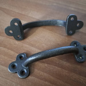May include: Two antique-style, dark gray metal drawer pulls. Each pull has a curved handle and decorative, clover-shaped mounting plates with three holes for screws. The pulls are designed for furniture and cabinets.
