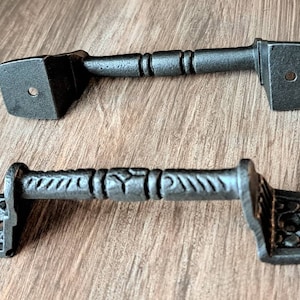 May include: Two black cast iron drawer pulls with ornate designs. The top pull has a simple design with a round knob on the end. The bottom pull has a more intricate design with a detailed pattern.