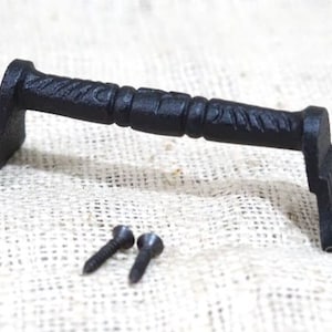 May include: Black cast iron handle with decorative detailing. The handle has a rectangular base with a floral design and a curved grip. Two black screws are included. Ideal for cabinets or drawers.