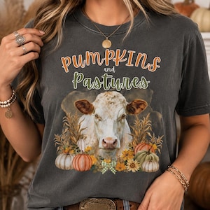 May include: A charcoal gray t-shirt featuring a graphic of a white cow surrounded by pumpkins, sunflowers, and dried wheat. The text "pumpkins and Pastures" is printed in orange and green above the cow illustration, which includes a small green bow.